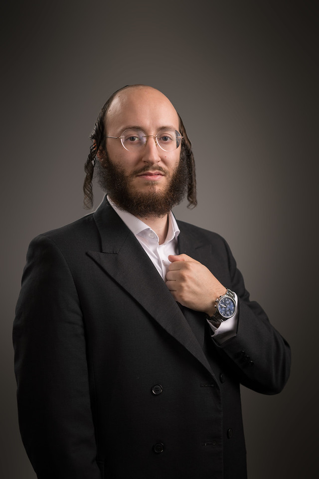 A man wearing glasses and a dark jacket holds his lapel, looking directly at the camera against a warm gray backdrop.