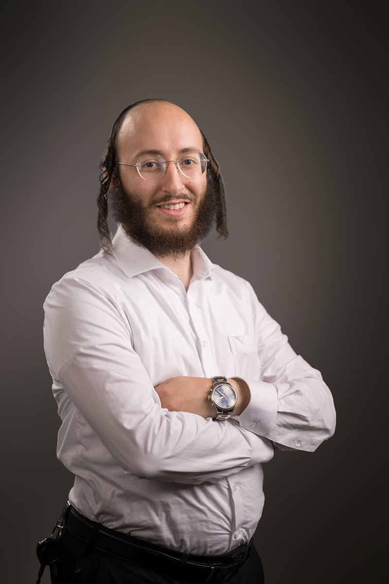 A man wearing glasses and a white shirt smiles with his arms crossed against a warm gray backdrop.