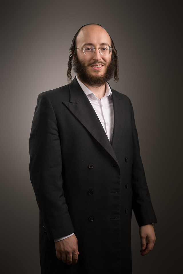 A man in a dark double-breasted suit jacket stands with his hands at his sides against a warm gray backdrop.