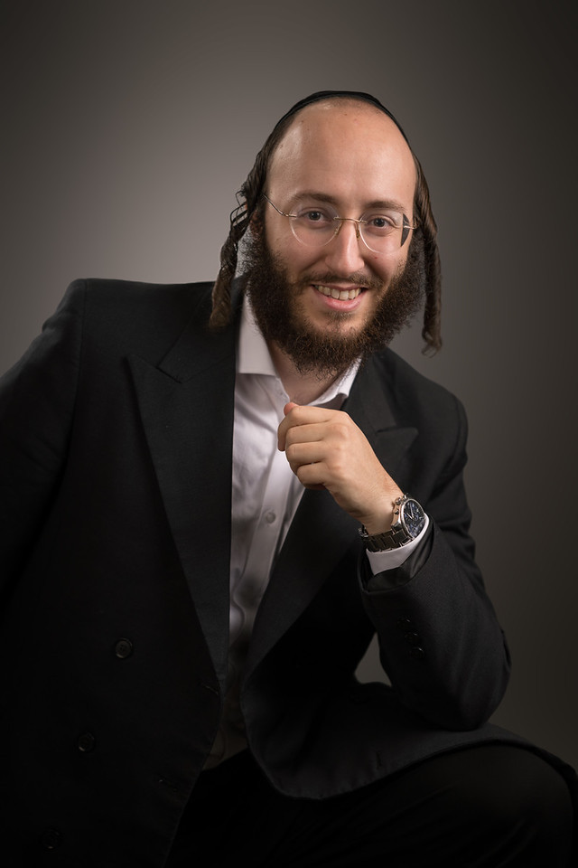 A smiling man in a dark jacket leans forward while seated, with one hand near his chin.